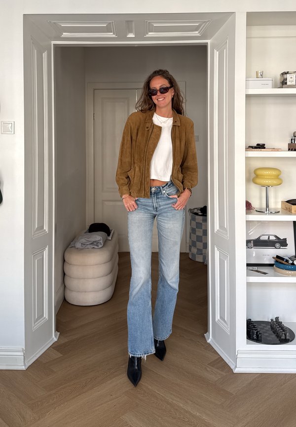 Una mujer con gafas de sol, chaqueta marrón, top blanco, vaqueros y botas negras posa en una puerta junto a unas estanterías con adornos.