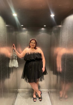 Vestido negro sin tirantes de tul con falda en capas, complementado con bolsos de lentejuelas. Las lisas paredes metálicas del ascensor reflejan la luz.