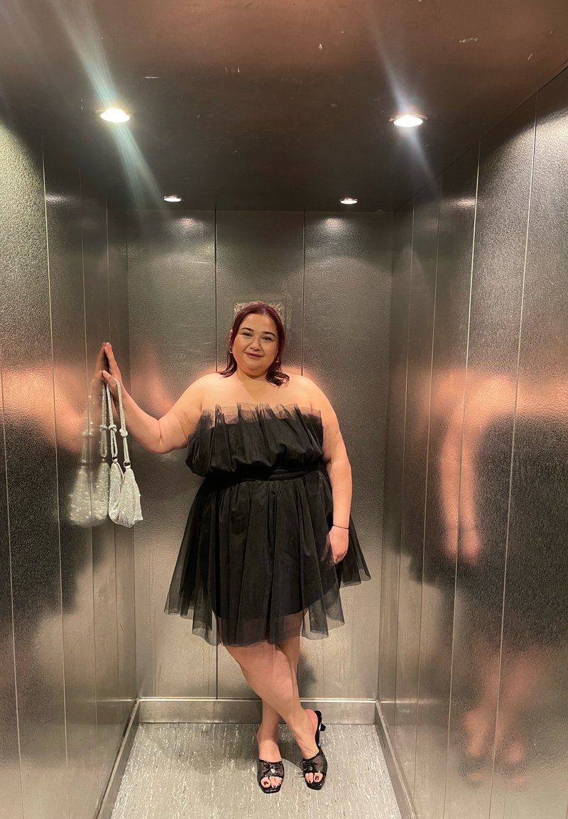 Robe noire en tulle sans bretelles avec une jupe à plusieurs couches, accessoirisée de sacs à main pailletés. Les murs métalliques lisses de l'ascenseur reflètent la lumière.