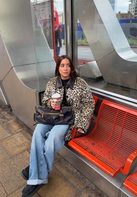 Jeune femme en manteau léopard tenant un café, assise sur un banc rouge à un arrêt de bus en métal avec une rue de la ville en arrière-plan.