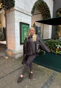 Przesadnie duża brązowa skórzana kurtka typu bomber noszona na ciemnym futerkowym topie, zestawiona z szarymi legginsami i brązowymi kapciami. W tle zielona witryna sklepu.