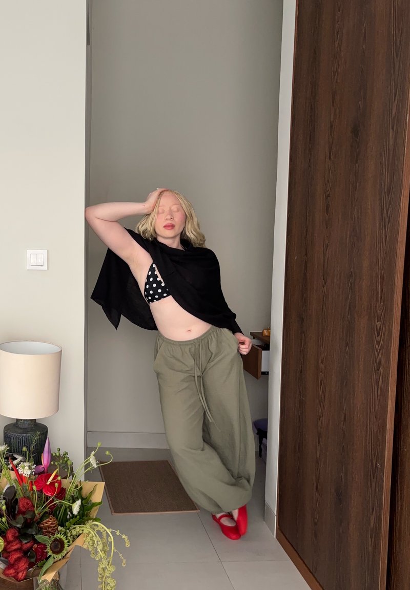 Person with light skin and blonde hair poses wearing a black top, polka dot bra, loose green pants, and red shoes in a hallway near flowers and a lamp.