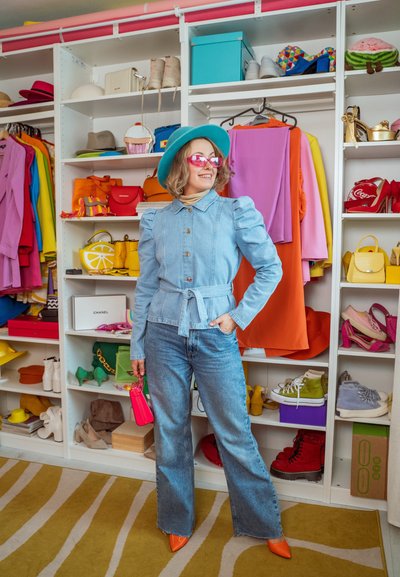 Chaqueta de mezclilla azul claro con mangas abullonadas y cintura ajustable, combinada con jeans acampanados. Los complementos incluyen un bolso rosa y tacones naranjas. Fondo de vestuario colorido.