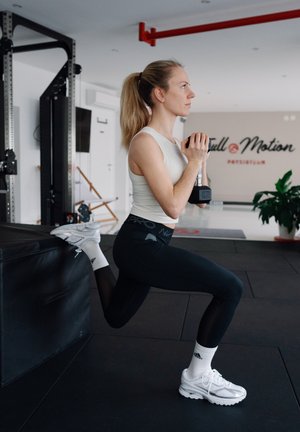 Una mujer realiza una zancada con una mancuerna en un gimnasio. Lleva una camiseta sin mangas, leggings negros con un patrón y zapatillas deportivas blancas.