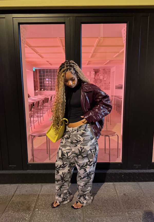 Jeune femme aux longs cheveux tressés portant un pantalon camouflage, une veste foncée et des sandales, se tient à l’extérieur d’un bâtiment vitré éclairé d’une lumière rose.