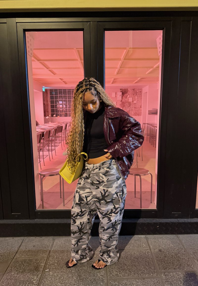 Jeune femme aux longs cheveux tressés portant un pantalon camouflage, une veste sombre et des sandales, se tient à l’extérieur d’un bâtiment vitré éclairé d’une lumière rose.