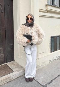 Plysjaktig beige kort jakke, hvite vide bukser, brun skjerf og liten svart veske. Mørke solbriller og brune ankelstøvler fullfører antrekket.