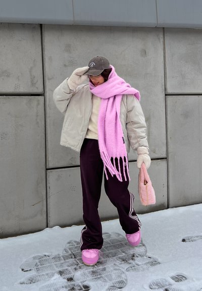 Chaqueta beige acolchada, suéter beige claro, pantalones de chándal morados oscuros con rayas blancas, pañuelo rosa con flecos, bolso rosa peludo, zapatos rosas. Fondo nevado.
