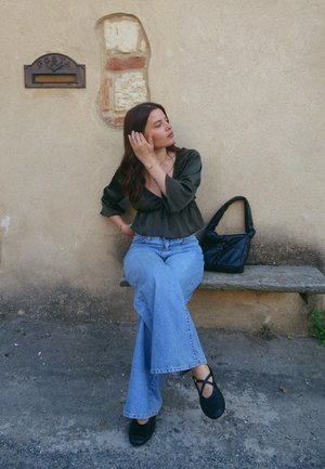 Outfit casual: blusa de manga larga color oliva, jeans de campana azul claro, bolso negro y zapatos negros. Sentada en un banco de piedra contra una pared texturizada.