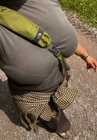 Person med grå skjorte og ruted skjorte knyttet rundt livet med en grønn North Face stropp over brystet, som står på en grusvei.