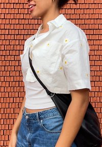 Valge lühike särk, millel on kollased karikakrad, on kantud kinnise valge kampsuni üle, koos siniste teksade ja musta risti kehakotiga.