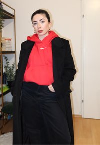 Un sweat à capuche rouge Nike sous un manteau noir ; le mannequin porte un jean foncé. La pièce comprend des étagères à livres et des plantes vertes en arrière-plan.