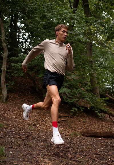 Corredor con una camiseta de manga larga beige claro y pantalones cortos negros, usando zapatillas blancas con toques rojos, navegando por un sendero boscosa.