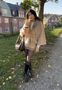 Beige kappa med pälskrage, vit tröja, hundtandsmönstrad kjol, svarta strumpbyxor och ankelstövlar. Håller en kaffe i en pappersmugg.