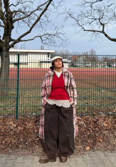 Joven con abrigo de cuadros y gorra, de pie junto a una valla metálica con suelo cubierto de hojas y árboles desnudos en una zona urbana al aire libre.