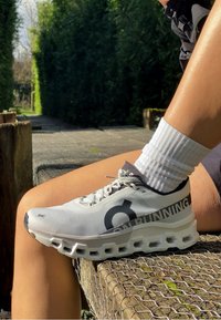 Fehér sneaker fekete részletekkel, lélegző hálós felsőrésszel, vastag párnázott talppal és "ON RUNNING" felirattal az oldalon. Fehér zoknival viselve.