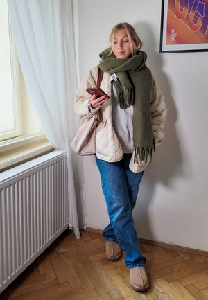 Chaqueta acolchada beige, sudadera gris, bufanda verde oliva, vaqueros azules y botas de tobillo color camel, sosteniendo un teléfono inteligente. Fondo claro con una ventana.