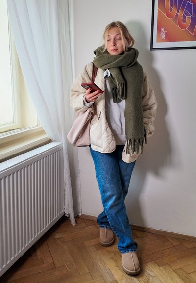 Chaqueta acolchada beige, sudadera gris, bufanda verde oliva, vaqueros azules y botas de tobillo color camel, sosteniendo un teléfono inteligente. Fondo claro con una ventana.