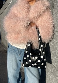 Personne portant une veste rose duveteuse et un jean bleu clair, tenant un sac à main noir à gros pois blancs, debout contre un mur gris.