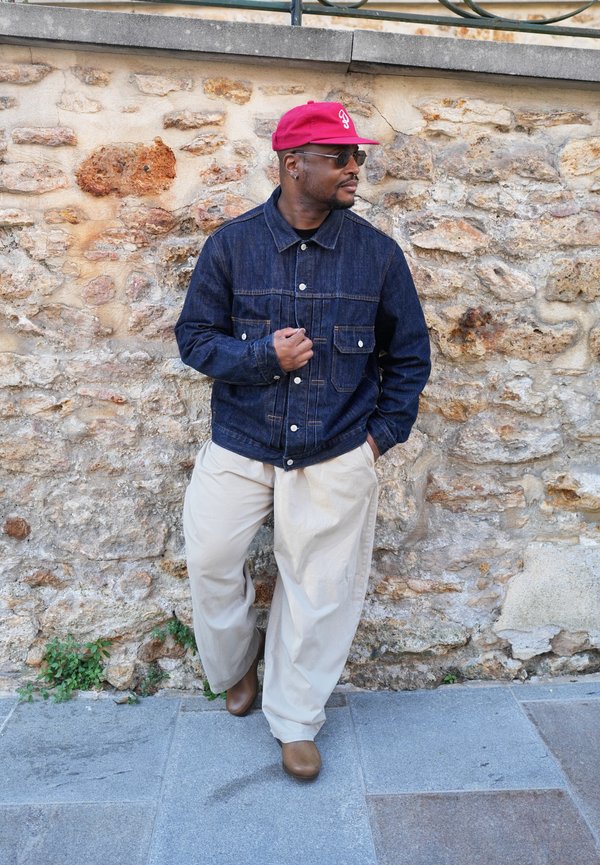 Uomo con berretto rosso, occhiali da sole, giacca di jeans scura, pantaloni beige larghi e scarpe marroni che si appoggia a un muro di pietra su un marciapiede.