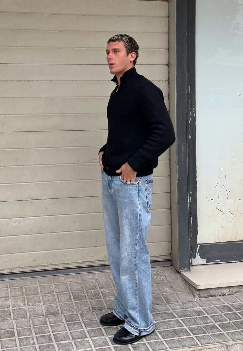 Jeune homme aux cheveux bouclés debout les mains dans les poches, portant un pull noir zippé, un jean bleu clair ample et des chaussures noires sur un trottoir carrelé.