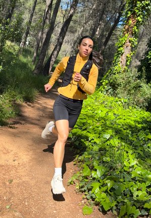 Atleta con una camiseta de manga larga amarilla brillante y pantalones cortos negros, que lleva un chaleco negro con bolsillos, corriendo por un sendero de tierra rodeado de vegetación.