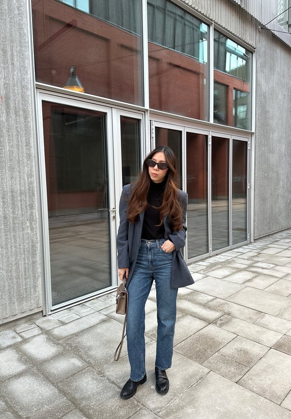 Blazer cinza sobre um pulôver de gola alta preto, jeans azuis de cintura alta, sapatos pretos, segurando uma pequena mala castanha, com portas de vidro ao fundo.