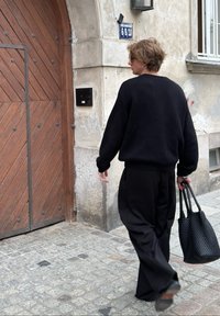 Zwarte oversized trui en losse zwarte broek, met een gestructureerde zwarte tas, wandelt langs een houten deur met een diagonaal patroon.
