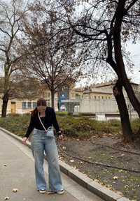 Φαρδιά ανοιχτόχρωμα μπλου τζιν, μαύρη μπλούζα με μακρύ μανίκι, μικρή ασημένια τσάντα, λευκά αθλητικά παπούτσια, στέκεται σε πλακόστρωτο πεζοδρόμιο με γυμνά δέντρα.