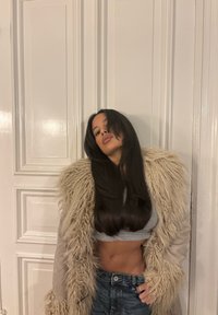 Manteau en fausse fourrure beige à texture ébouriffée, haut gris court, et jean en denim déchiré. Longs cheveux foncés partiellement couvrant le visage.