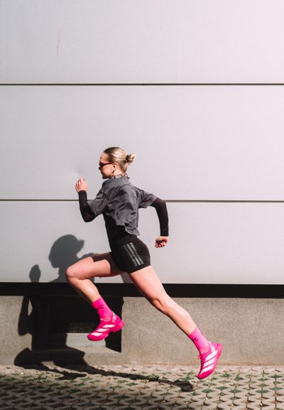Atleta con gafas de sol, camiseta gris, pantalones cortos negros, y zapatos y calcetines de un rosa brillante corre a paso medio junto a una pared moderna gris al aire libre.