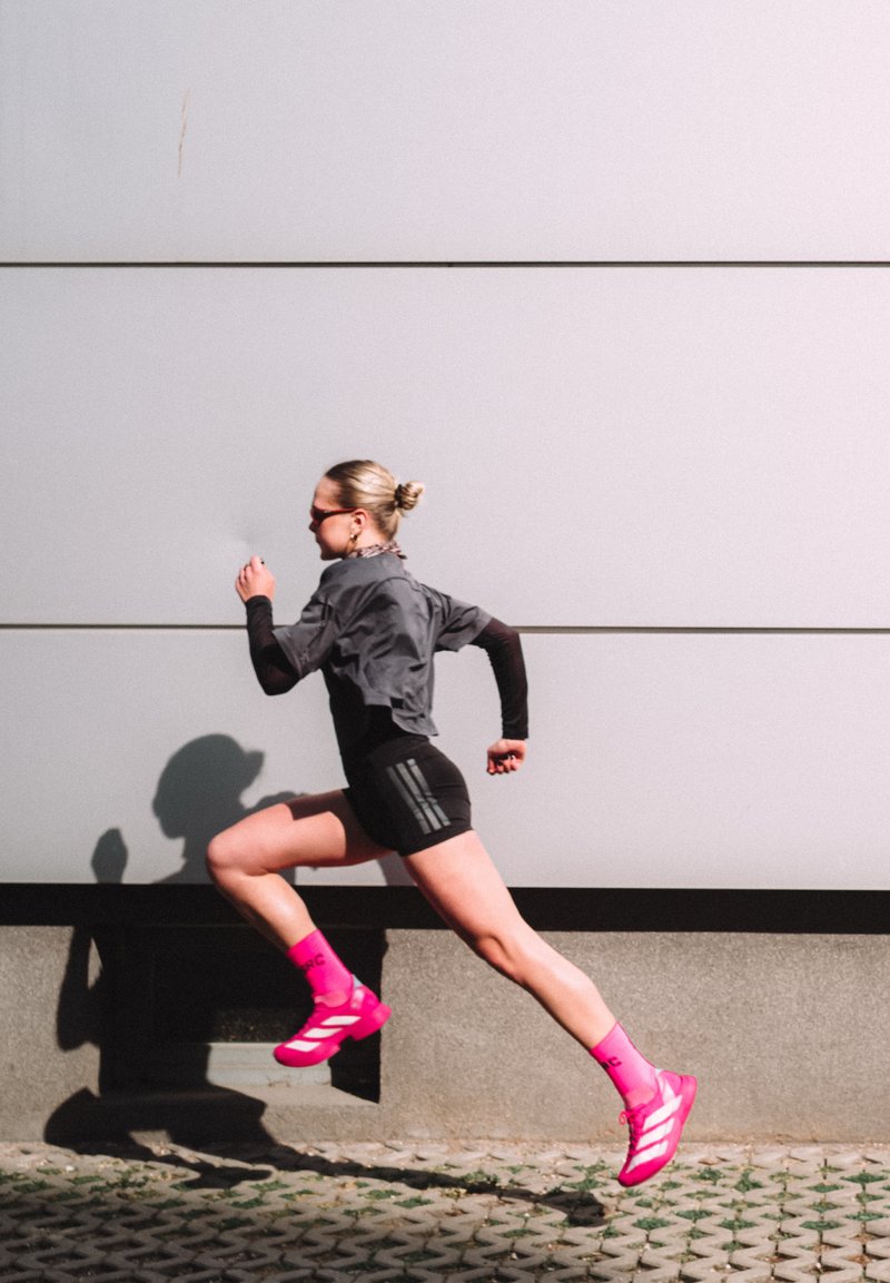 Αθλητής με γυαλιά ηλίου, γκρι μπλουζάκι, μαύρα σορτς και φωτεινά ροζ παπούτσια και κάλτσες τρέχει μέτριο βήμα δίπλα σε έναν μοντέρνο γκρι τοίχο έξω.