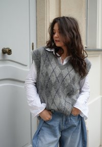 Grijze gebreide vest met een ruitpatroon, gedragen over een wit shirt, gecombineerd met lichtblauwe denim broek. Zachte textuur en losse pasvorm.