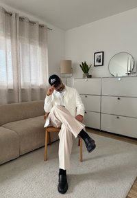 Fehér blézert, bézs nadrágot és fekete sapkát viselő férfi egy fa széken ül egy modern nappaliban, ahol bézs kanapé, szekrény, lámpa és növény található.