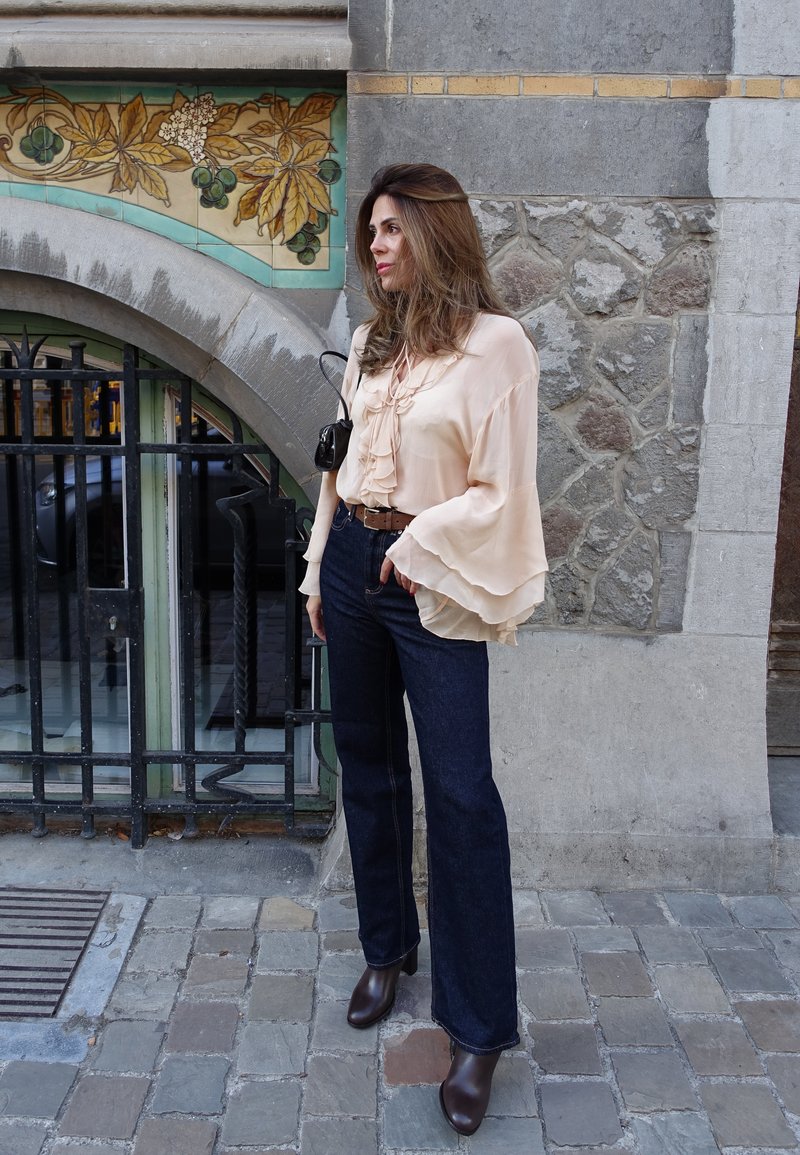 Blusa de pêssego claro, com babados e mangas largas, combinada com jeans azuis escuros e boca de sino e botas de tornozelo castanhas. Acessório: pequena bolsa preta.