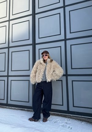 Mujer con chaqueta beige de peluche, vaqueros oscuros de pata ancha, zapatos marrones, gafas de sol y gorra marrón, de pie sobre la nieve frente a una gran puerta panelada.