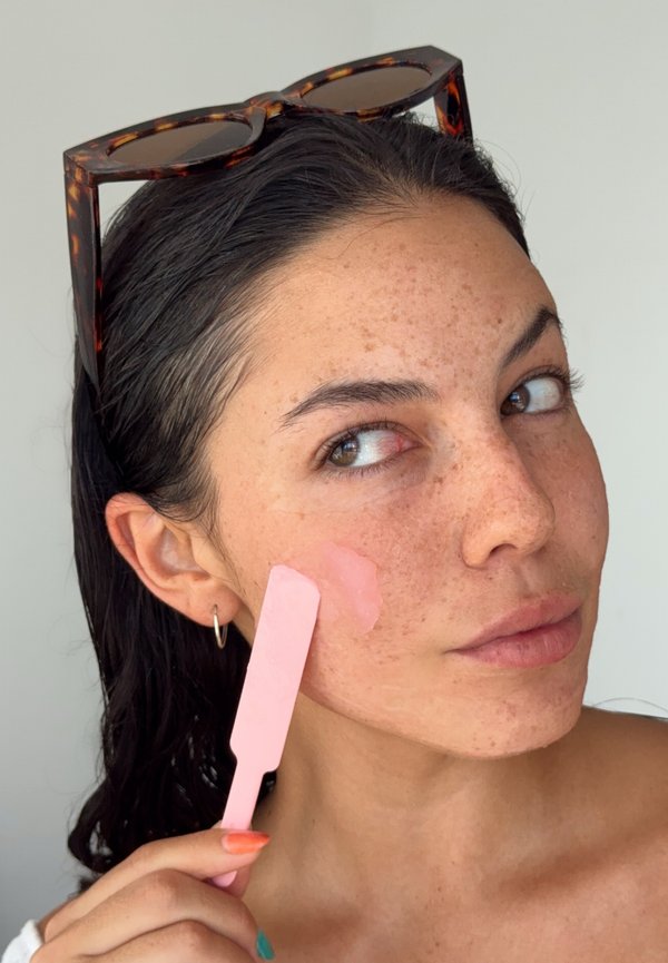 Donna con lentiggini che applica crema rosa sulla guancia usando una spatola, occhiali da sole sulla testa, guarda di lato.