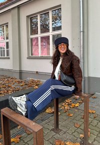 Kvinde med langt hår iført mønstret jakke, orangefarvede briller, sort baret, blåstribede bukser og hvide sneakers, der sidder på et metal cykelstativ udendørs.