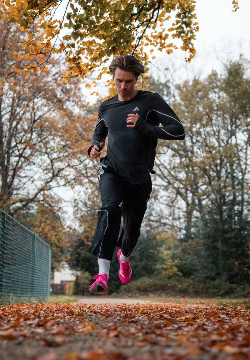 Man in black sportswear and bright pink running shoes jogging on a leaf-covered path in an autumn park.