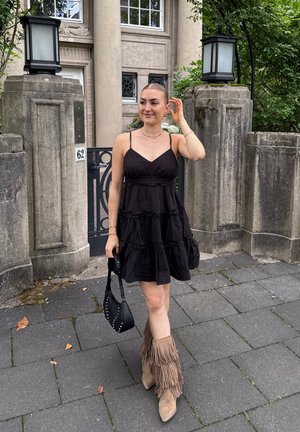 Mujer con un vestido negro sin mangas, sosteniendo un bolso negro, vistiendo botas de flecos beige, de pie sobre un camino de piedra fuera de un edificio de piedra con verja.