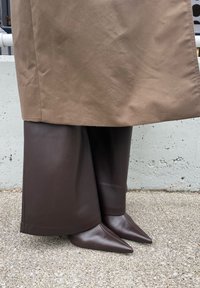 Hnedé kožené rozšírené nohavice s hladkou textúrou, doplnené hnedými lodičkami na špicatú špičku. Komplekt dopĺňa dlhý hnedý kabát.