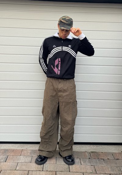 Persona con una gorra de camuflaje, chaqueta deportiva negra de Adidas, pantalones cargo marrones sueltos y zapatos negros robustos, de pie contra una pared blanca.