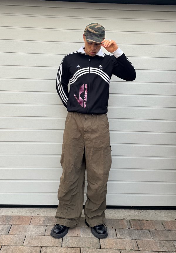 Osoba w kamuflażowej czapce z daszkiem, czarnej sportowej kurtce Adidas, luźnych brązowych spodniach typu cargo i czarnych masywnych butach stojąca na tle białej ściany.