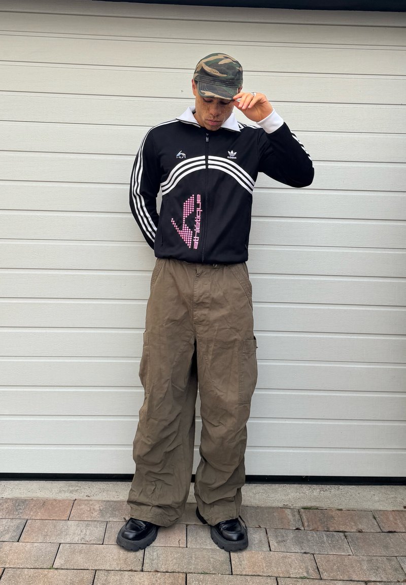 Άτομο που φοράει καπέλο παραλλαγής, μαύρο μπουφάν Adidas, φαρδιά καφέ παντελόνια cargo και μαύρα ογκώδη παπούτσια, στέκεται σε λευκό τοίχο.