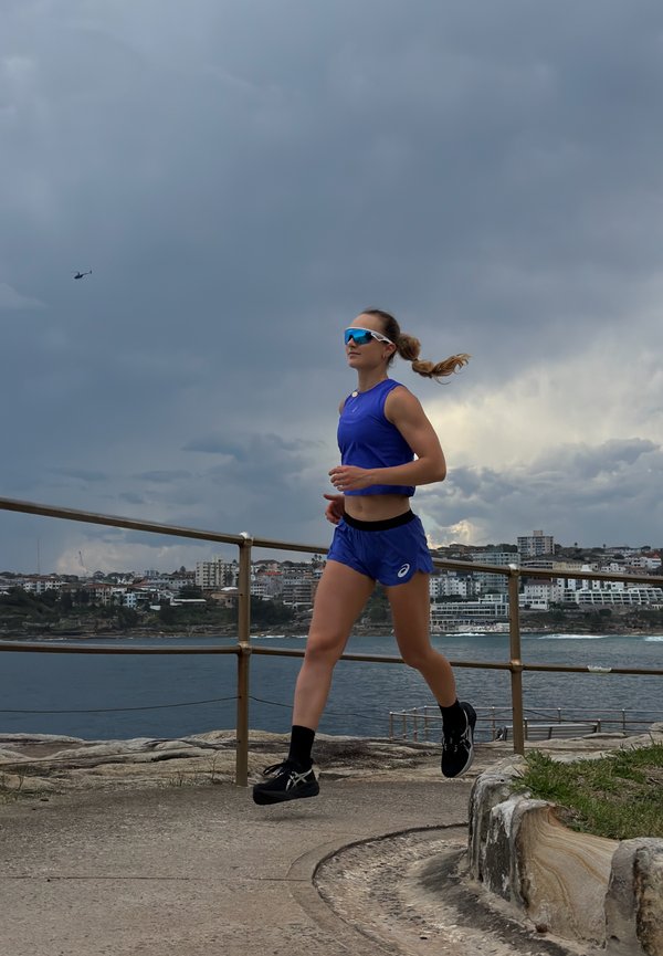 Femeie atletă în echipament de alergare albastru și ochelari de soare jogging pe o potecă de pe litoral cu cer înnorat și clădiri ale orașului în depărtare.