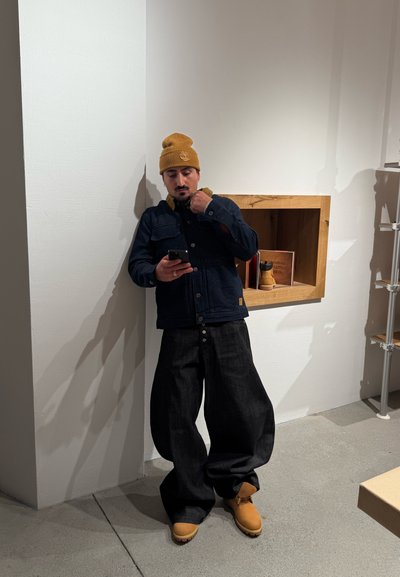 Hombre con gorro mostaza, chaqueta oscura, pantalones anchos negros y botas marrones, de pie en el interior y mirando un teléfono inteligente.