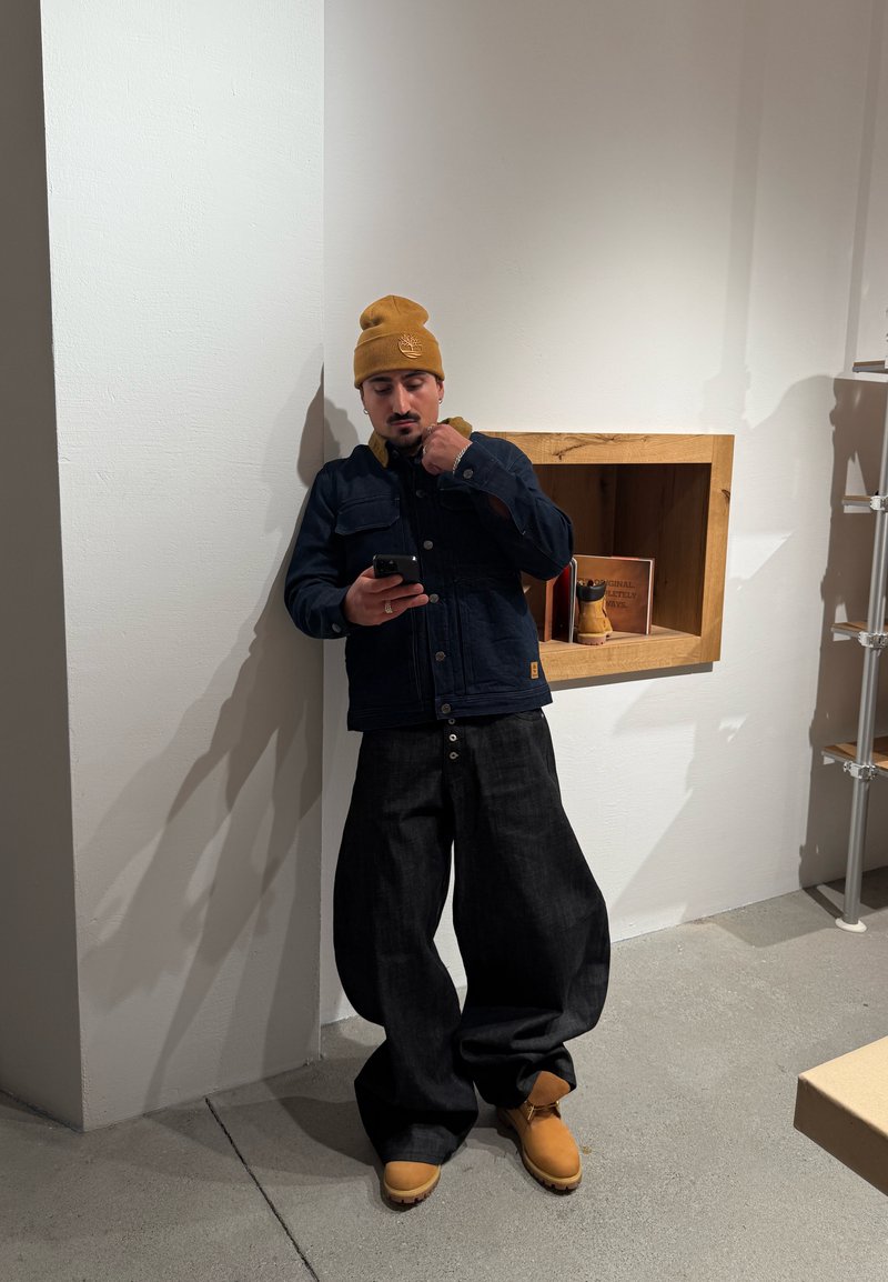 Homme portant un bonnet moutarde, une veste foncée, un pantalon large noir et des bottes marron clair, debout à l'intérieur en regardant un smartphone.