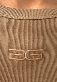 Gros plan sur l'encolure côtelée d'un pull beige avec un logo géométrique beige brodé brillant centré sous le col.
