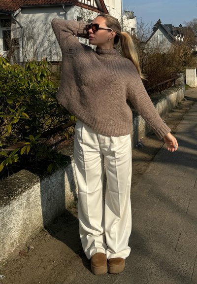 Suéter de punto texturizado marrón con cuello alto, combinado con pantalones blancos de pierna ancha y zapatos de plataforma color crema. Pose de pie al aire libre.