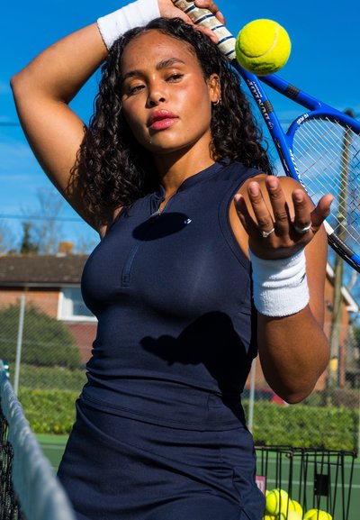 Vestido de tenis sin mangas en color azul marino con cuello con cremallera, tejido texturizado, combinado con muñequeras blancas. Se encuentran presentes una raqueta de tenis y una pelota.